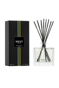 A reed diffuser bottle with black reeds next to its black packaging box, both featuring the NEST New York logo and a vertical blue and yellow stripe design, set against a plain white background.
