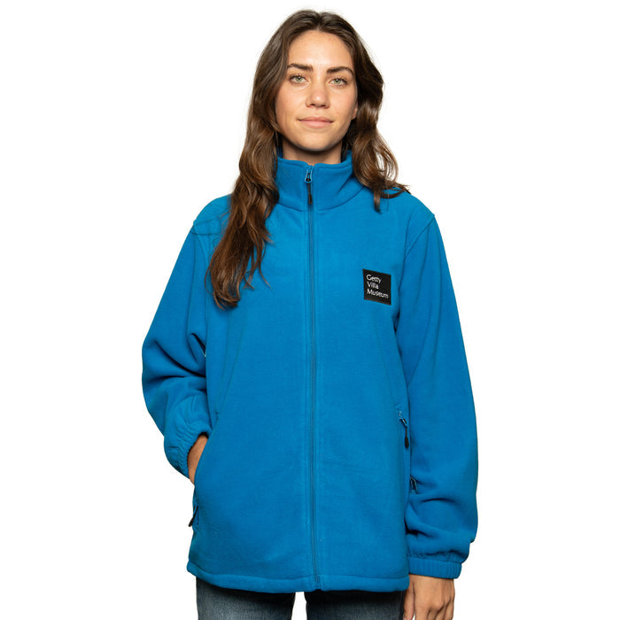 A woman wearing a blue full-zip fleece jacket with the Getty Villa Museum logo embroidered on the chest, standing against a plain background.