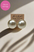 A pair of round, pearl-like clip-on earrings in a warm off-white/ivory shade, displayed on a small beige card labeled 'COUTURE COLLECTION'. The image is styled with dramatic, directional shadows on a light beige background, and a pink circular graphic overlay in the top-left corner reading 'Clip-on Earrings'.